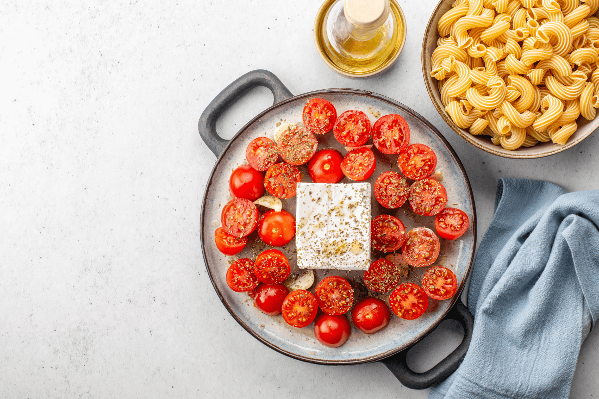 Gericht mit Feta-Käse und Tomaten, gewürzt, in einer Auflaufform, daneben gekochte Pasta und Olivenöl.