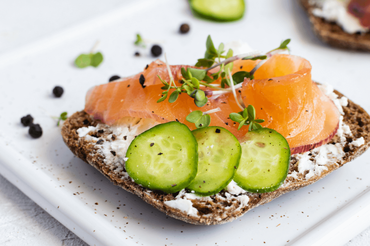 Belegtes Brot mit Frischkäse, Räucherlachs und Gurkenscheiben, garniert mit Sprossen und Pfefferkörnern auf weißem Teller.