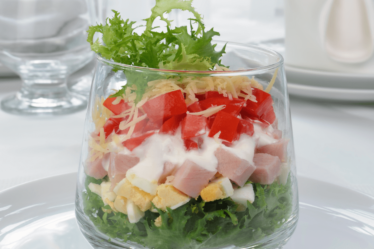 Schichtsalat im Glas mit grünem Blattgemüse, gewürfeltem Schinken, Tomaten, Käse und Dressing.