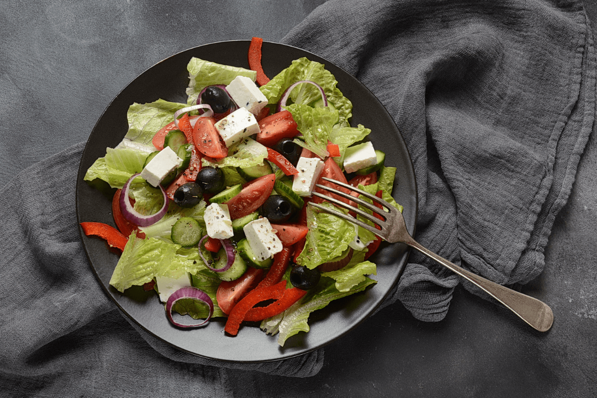 Schwarzer Teller mit griechischem Salat: Kopfsalat, Tomaten, Gurken, Paprika, Oliven und Feta-Käse, garniert mit Zwiebeln und Pfeffer.