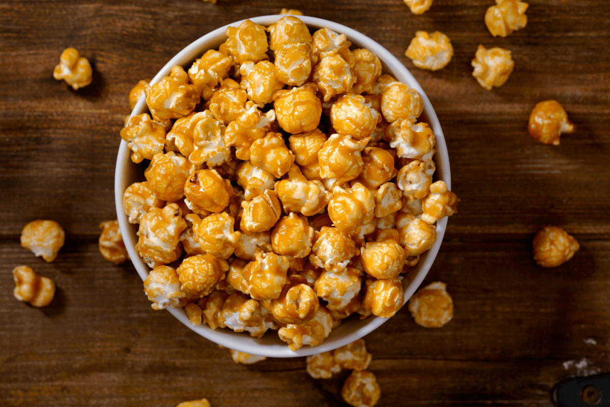 Schüssel mit karamellisiertem Popcorn, umgeben von einzelnen Karamel-Popcorn-Stücken auf einem Holzuntergrund.
