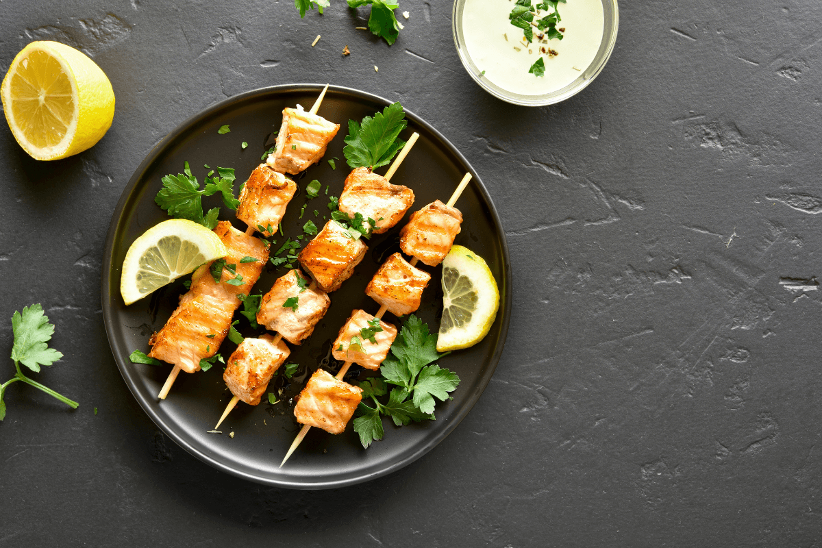 Gegrillte Lachs-Spieße auf schwarzem Teller, garniert mit Zitronenscheiben und frischer Petersilie.