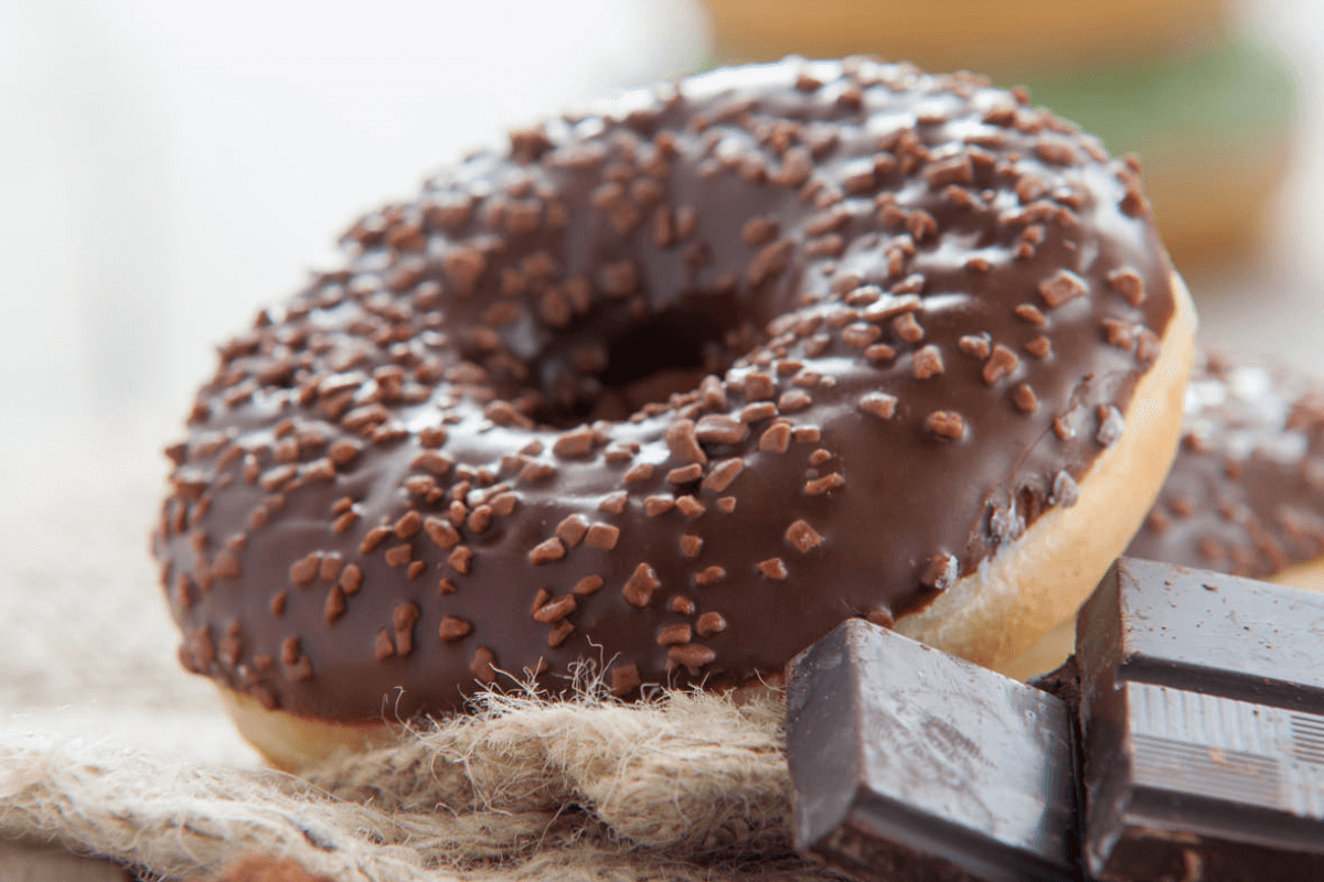 Schokoladenglasierte Mini-Donuts mit Schokostreuseln, neben einem Stück Zartbitterschokolade auf einem rustikalen Untergrund.
