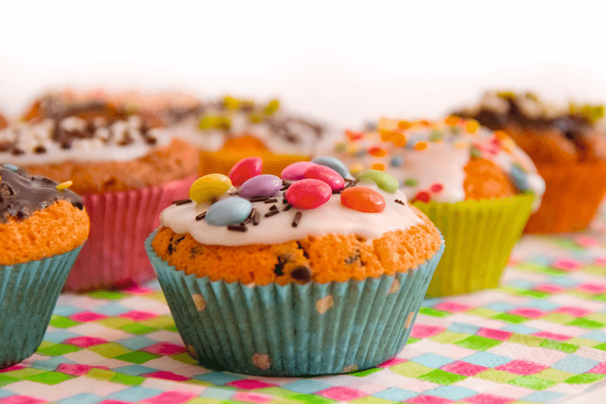Bunte Mini-Muffins in Papierförmchen mit Zuckerguss und bunten Streuseln auf einem karierten Tisch.