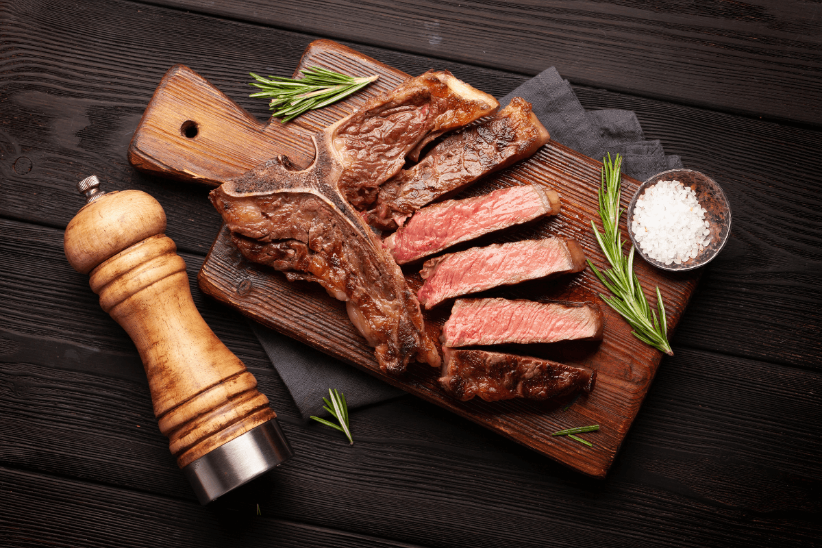 T-Bone Steak auf einem Holzbrett, begleitet von Rosmarin, grobem Salz und einer Pfeffermühle.