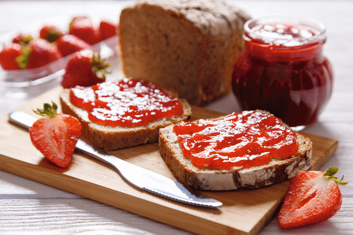 Zwei Scheiben Vollkornbrot mit Erdbeermarmelade, frische Erdbeeren und ein Glas Marmelade auf einem Holzbrett.