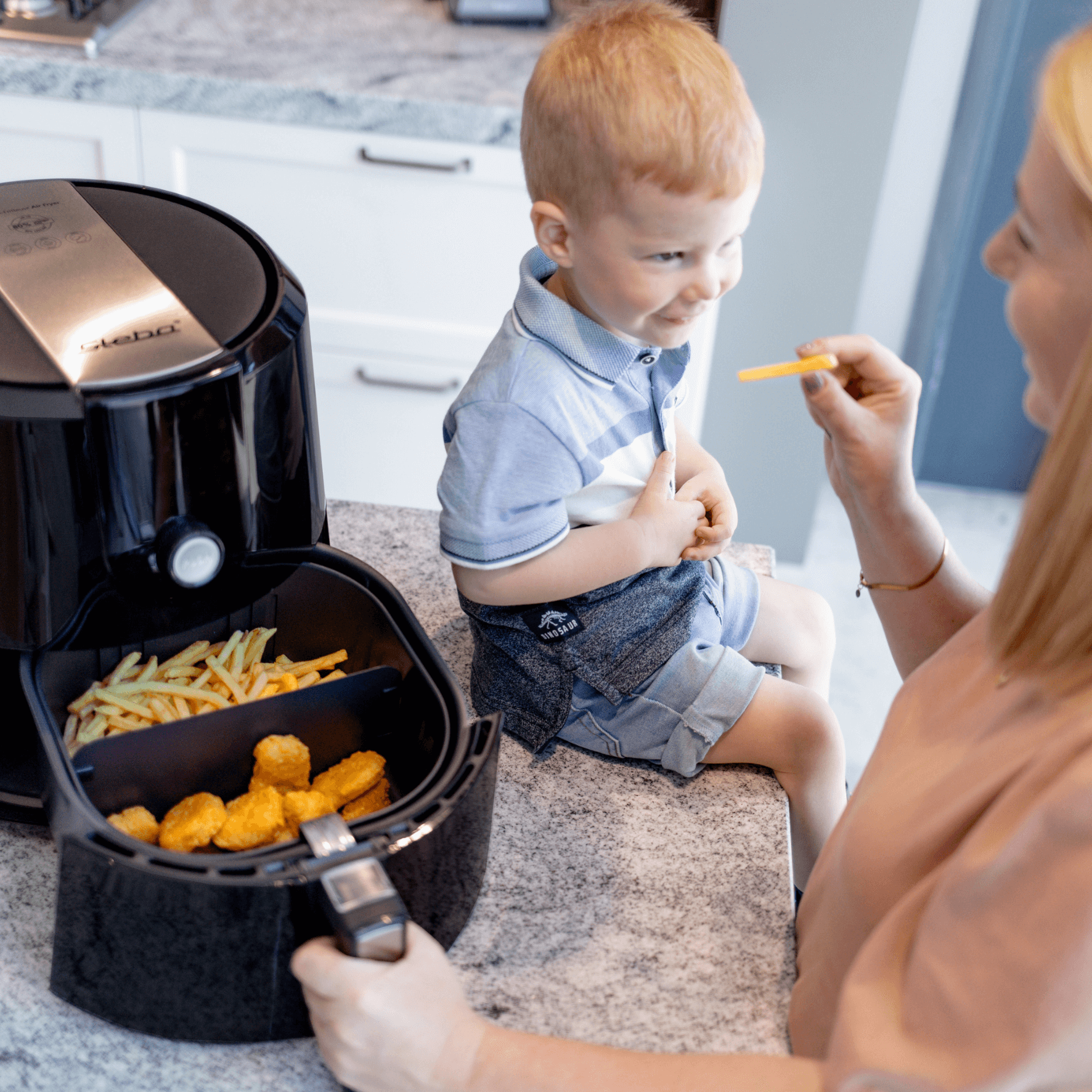 Heißluftfritteuse Steba HF5000 mit Pommes und Nuggets im Korb, daneben ein lächelndes Kind und eine Person, die ihm füttert.