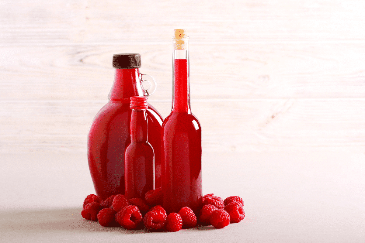 Drei Flaschen Himbeer Balsamico Essig in verschiedenen Größen, umgeben von frischen Himbeeren auf einem neutralen Untergrund.