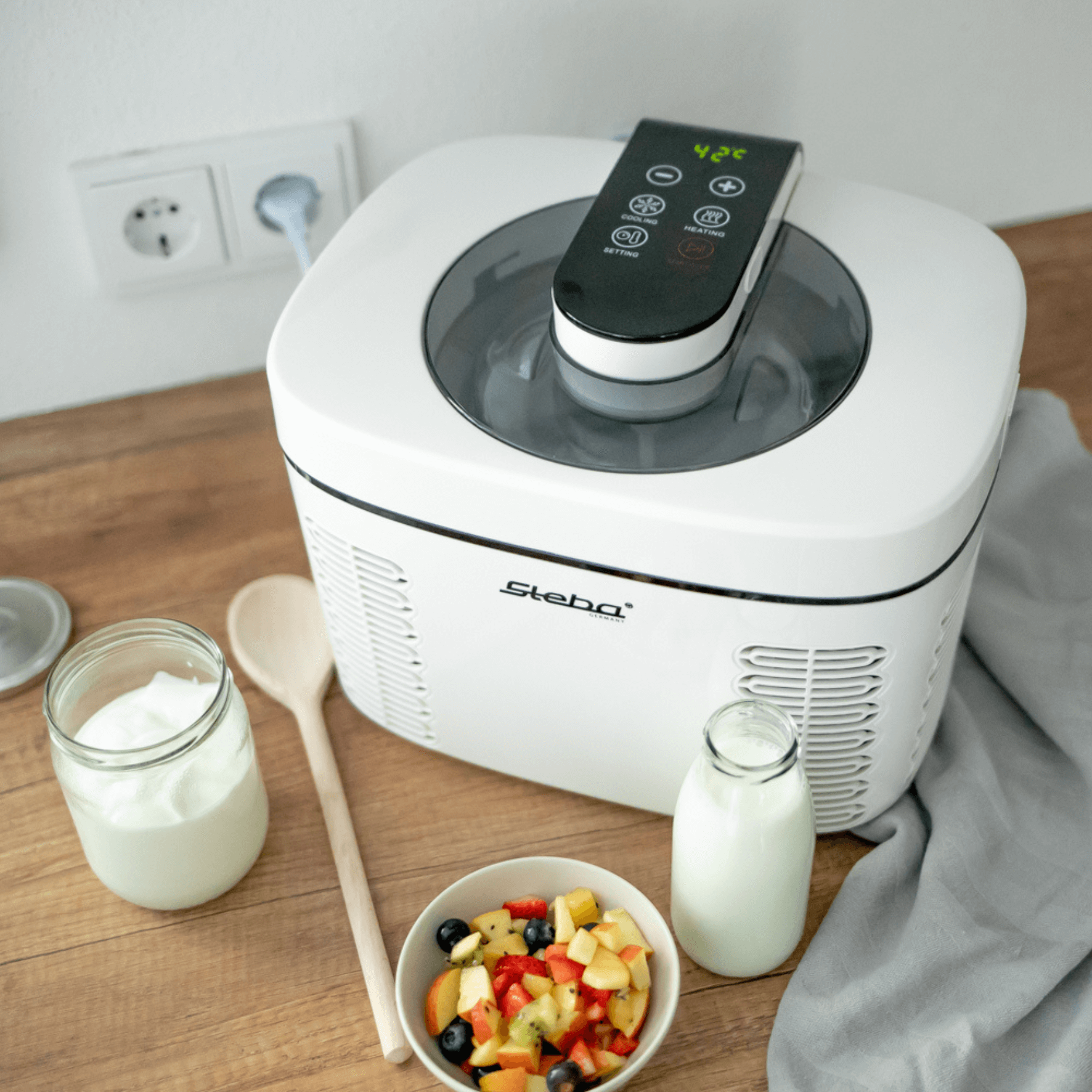Steba Eismaschine IC110 mit digitaler Anzeige, daneben frisches Obst, Joghurt und ein Holzlöffel auf einem Holzuntergrund.