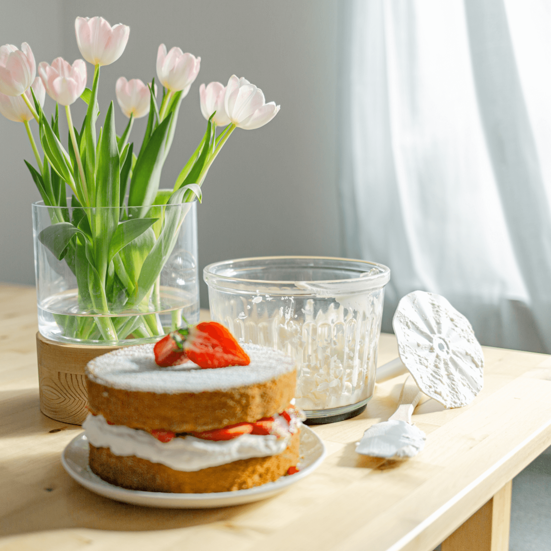 Ein frisch gebackener Erdbeerkuchen mit Sahne, dekoriert mit Erdbeeren, auf einem Tisch neben einem Glas mit Tulpen und einer Schüssel.