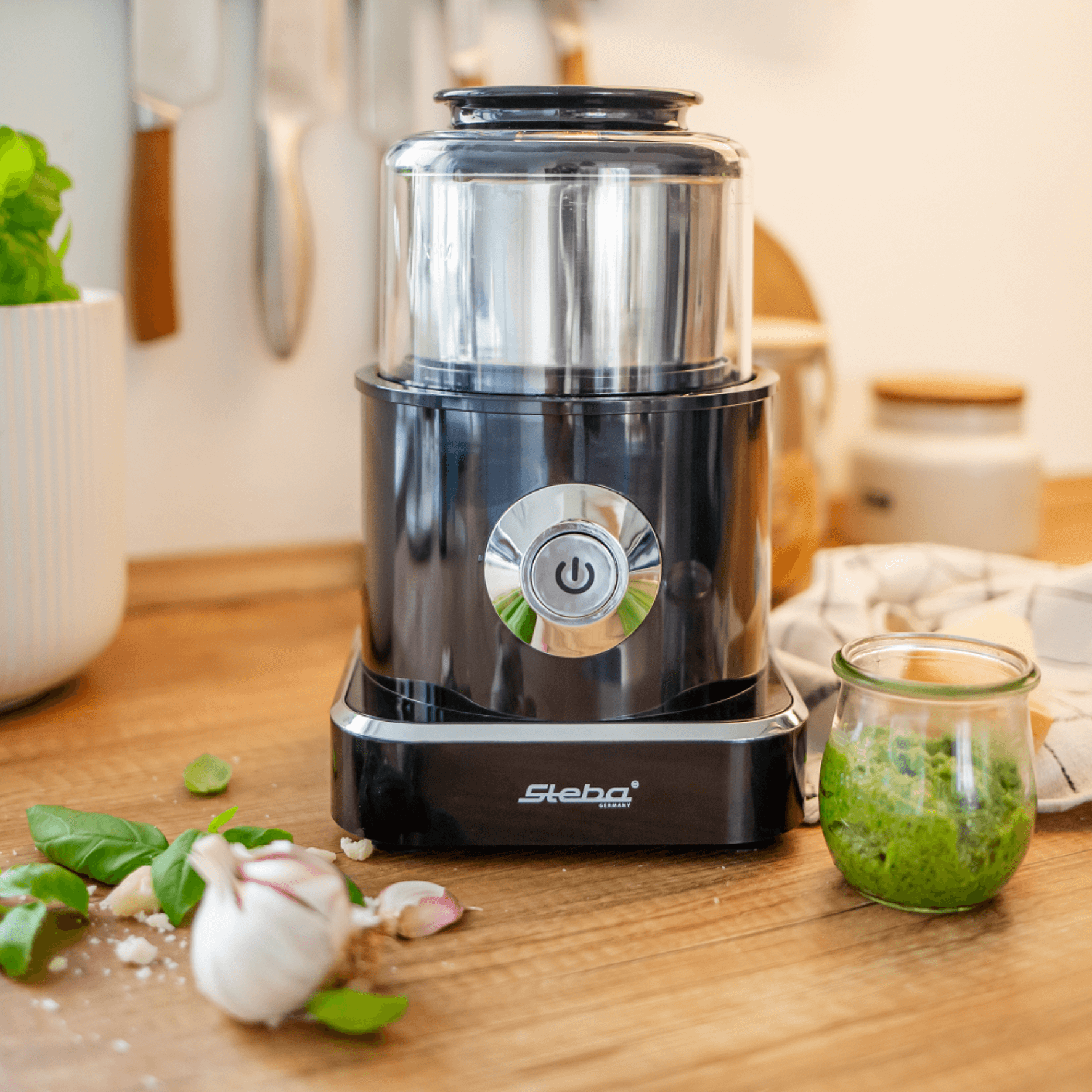 Steba Küchengerät in Schwarz mit Edelstahlbehälter, ideal zum Zerkleinern von Zutaten, umgeben von frischen Kräutern und einem Glas Pesto.