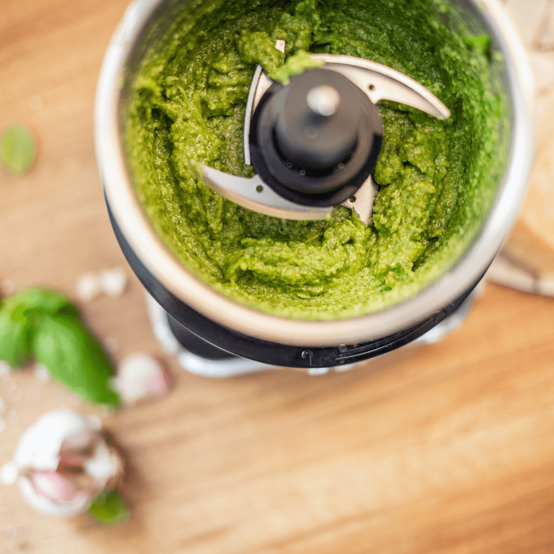 Grüner Pesto in einem Standmixer mit scharfen Klingen, umgeben von frischem Basilikum und Knoblauchzehen auf einem Holzuntergrund.
