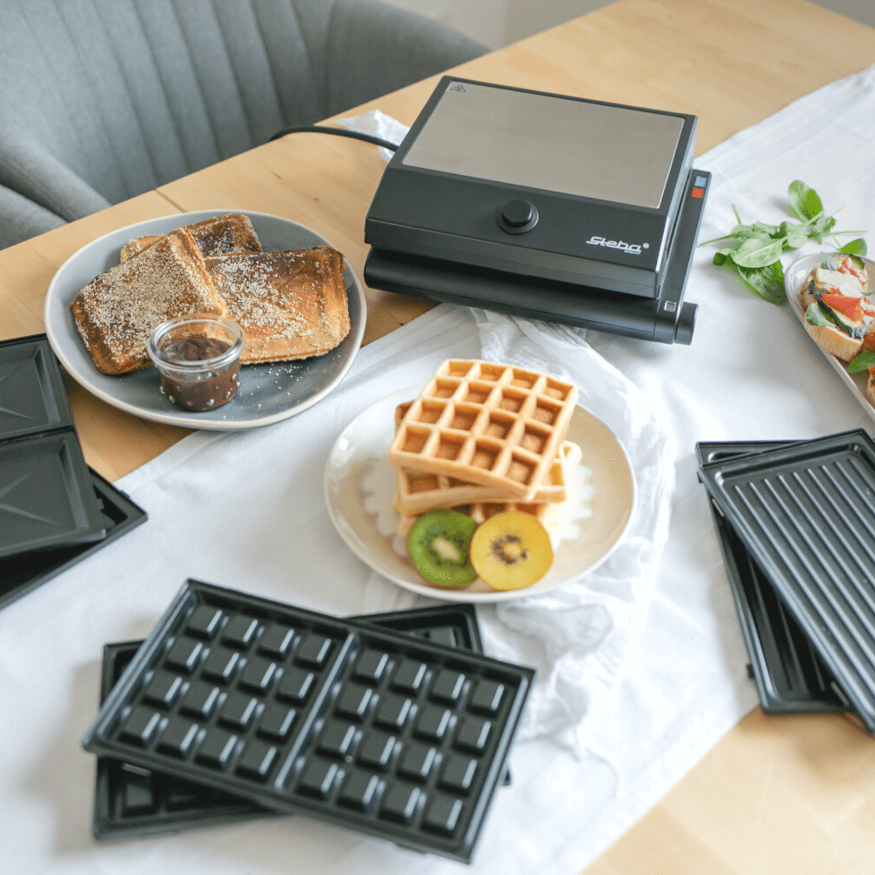 Steba SG55 Multi-Snack Maker mit Waffel- und Sandwichformen, auf einem Tisch mit Snacks und Obst.