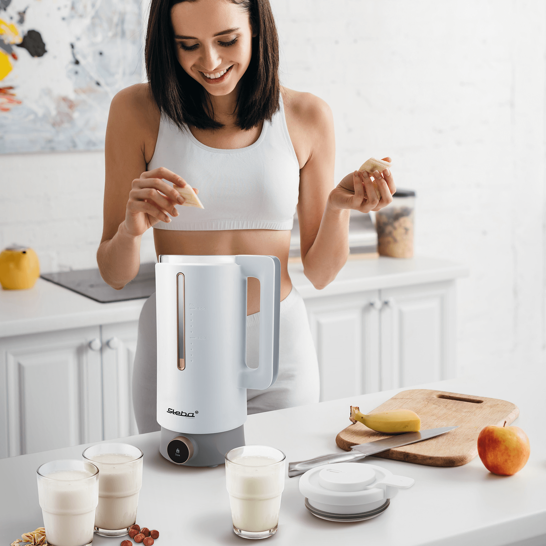 Vegan Drink Maker von Steba, weiße Farbe, mit transparentem Behälter. Person fügt Zutaten hinzu, drei Gläser mit Getränk im Vordergrund.