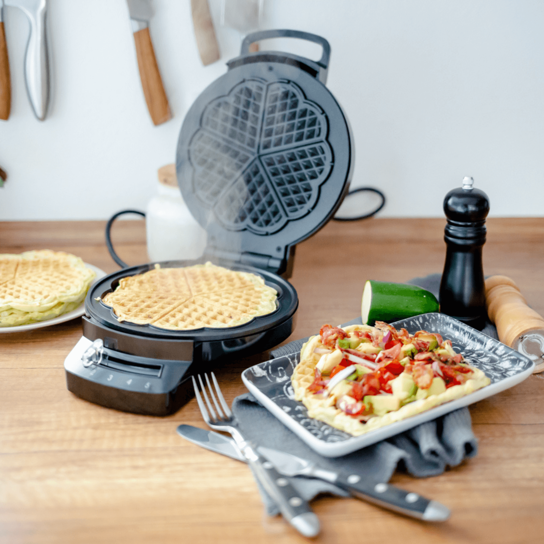 Waffeleisen von Steba mit dampfendem Waffelteig. Auf dem Tisch liegen bereits fertige Waffeln und ein Gemüsegericht.