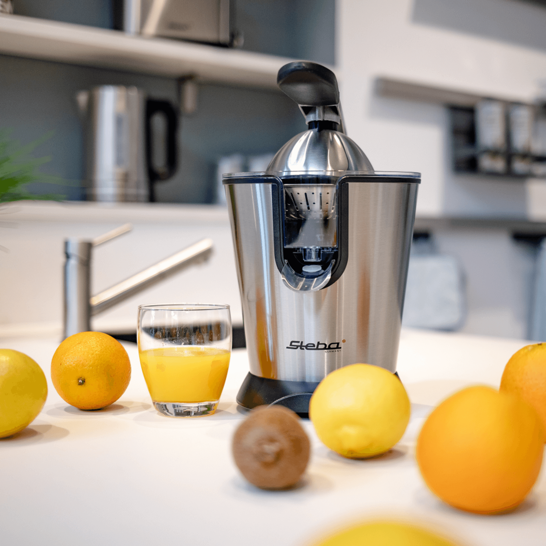 Steba Entsafter in Edelstahloptik, umgeben von frischen Zitrusfrüchten und einem Glas frisch gepresstem Saft auf einem Tisch.