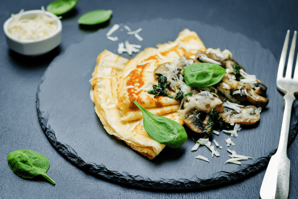 Pilz-Crêpe mit Käse auf schwarzem Teller, garniert mit frischem Spinat und geriebenem Käse.