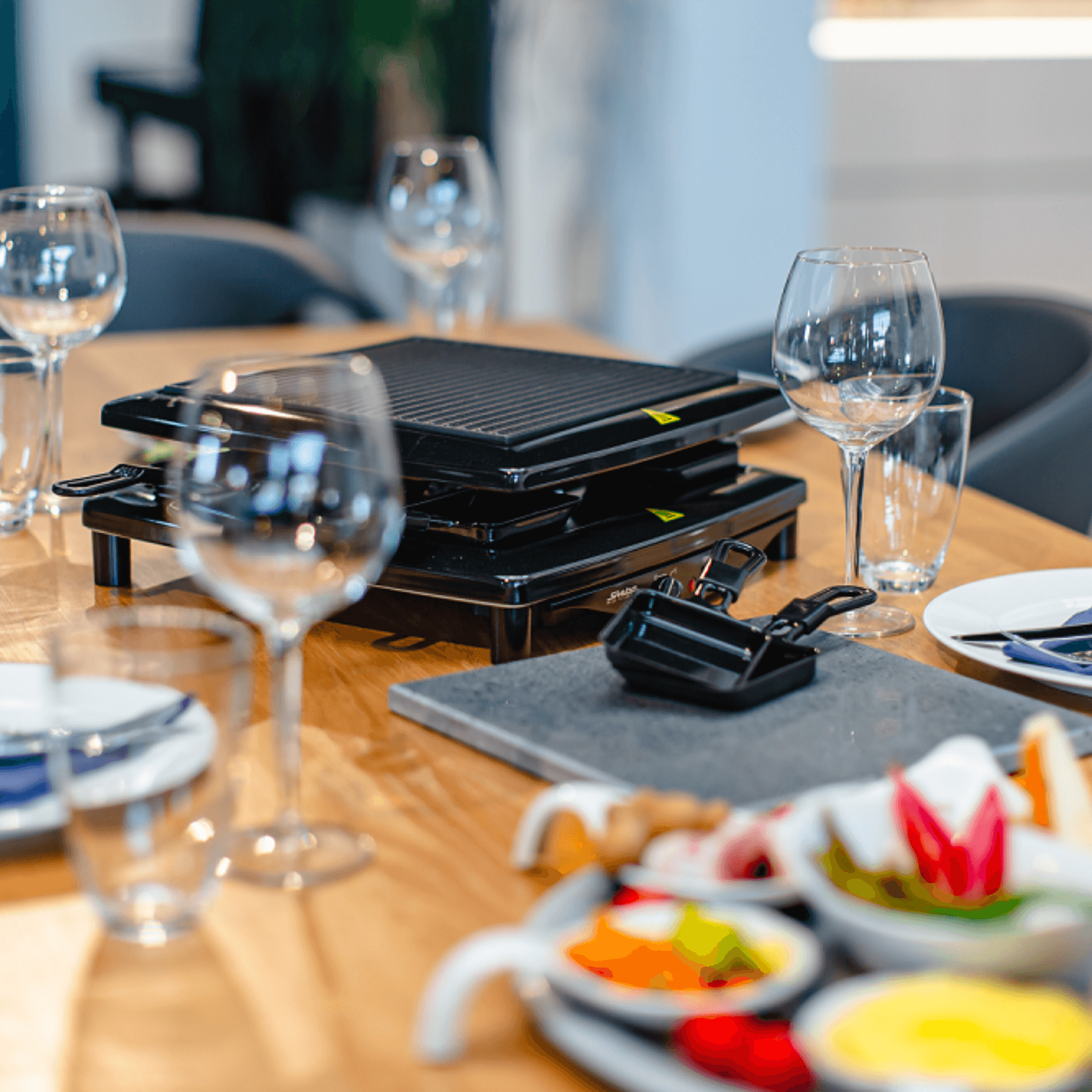 Steba Raclette-Grill RC3PLUS auf einem Tisch, umgeben von Gläsern und einer Auswahl an frischem Gemüse und Saucen.