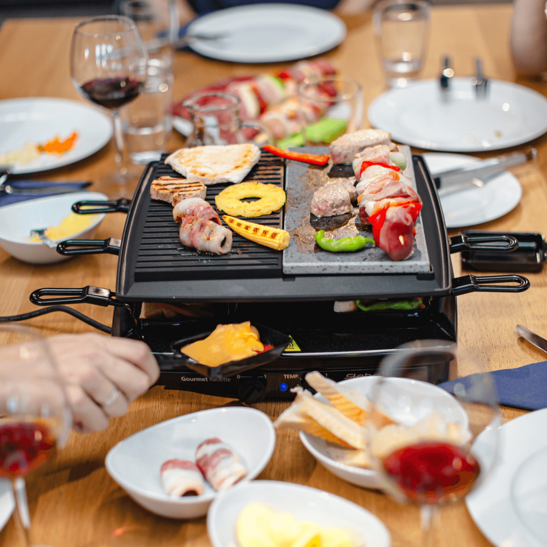 Raclette-Grill von Steba mit Grillfläche, verschiedene Speisen wie Gemüse und Fleisch, Gläser und Teller im Hintergrund.
