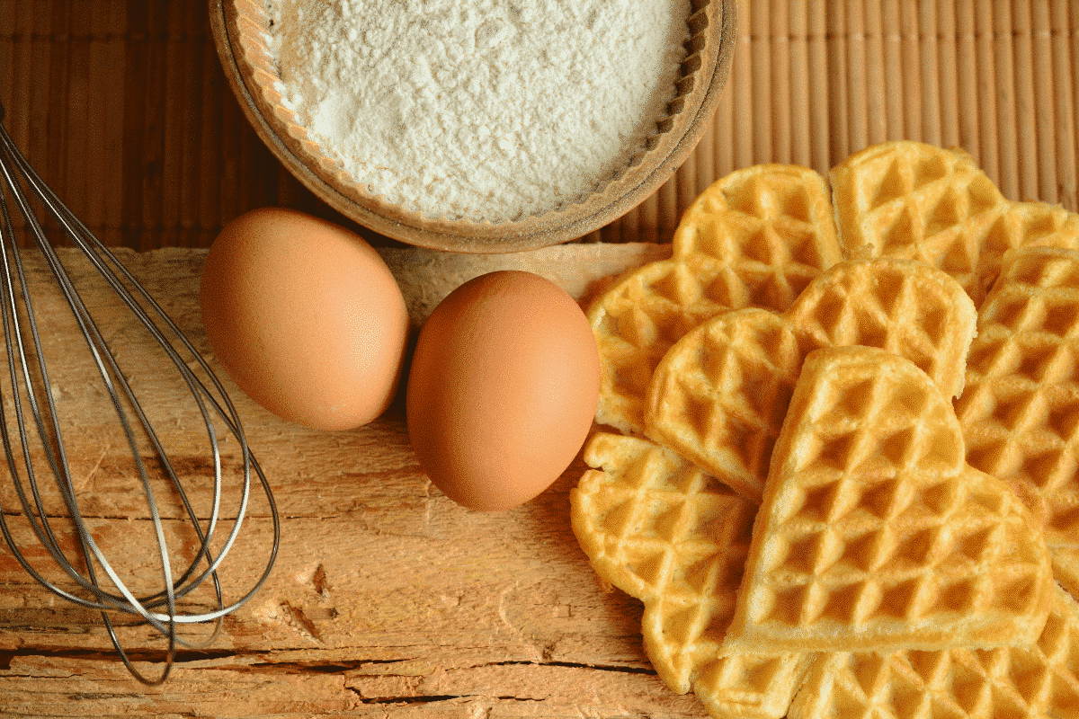 Zutaten für süße Waffeln: zwei Eier, Mehl in einer Schüssel und herzförmige Waffeln auf einem Holzbrett.