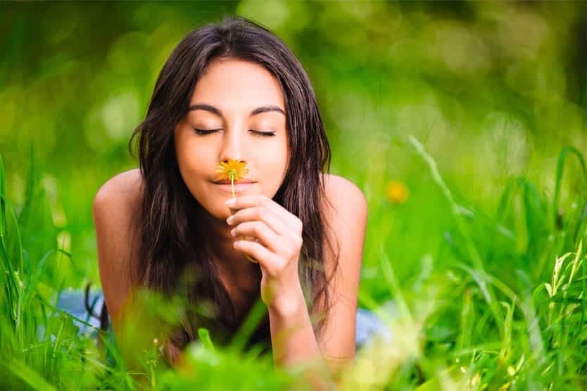Junge Frau liegt im Gras und hält eine Blume an die Nase, umgeben von grüner Natur.