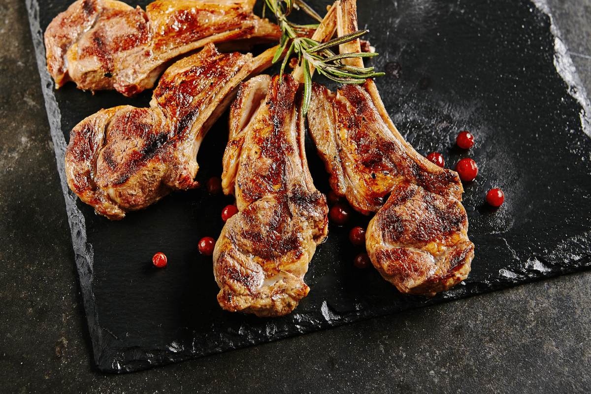 Marinierte Lammkoteletts auf einem schwarzen Steinbrett, garniert mit Rosmarin und roten Beeren.