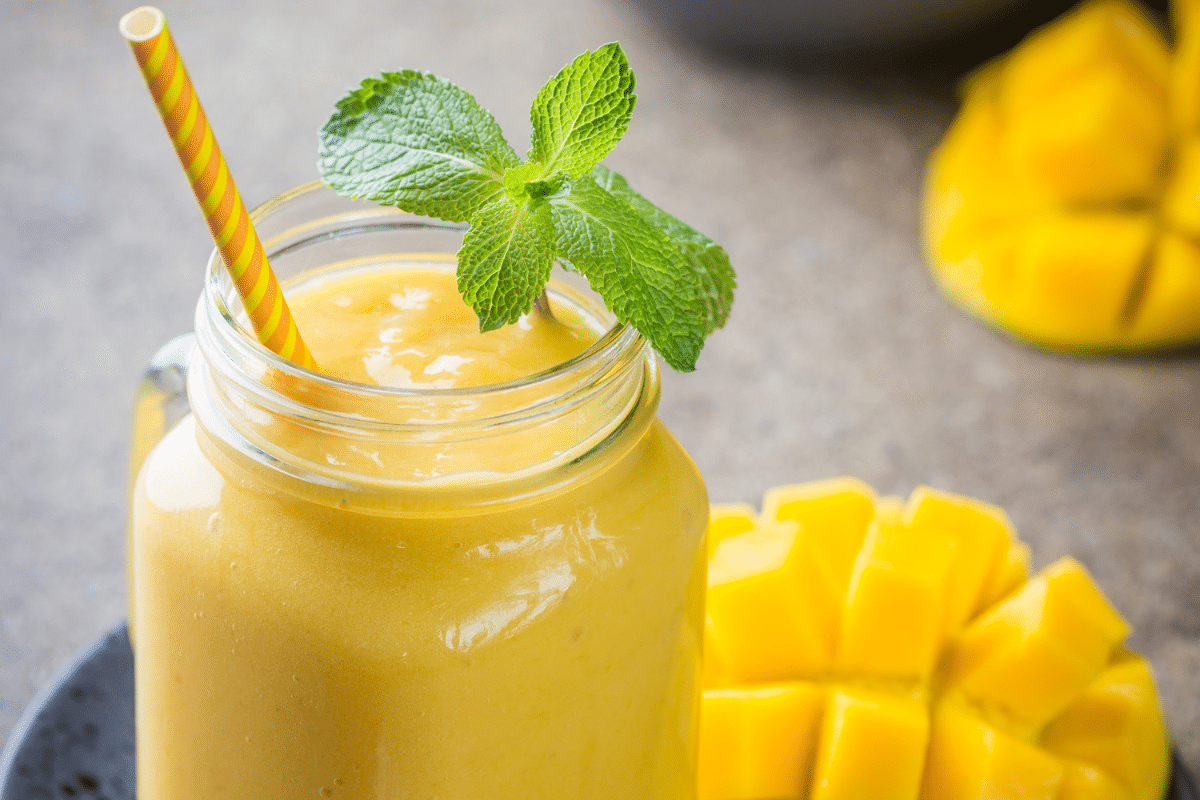 Ein erfrischender Sommer-Smoothie in einem Glas mit einem grünen Minzblatt und einem gestreiften Strohhalm, umgeben von frischen Mangostücken.