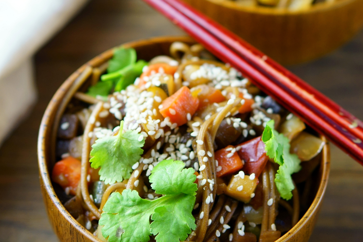 Eine Schale mit Glasnudelsalat, garniert mit Koriander, Sesam und buntem Gemüse, auf einem Holzuntergrund.