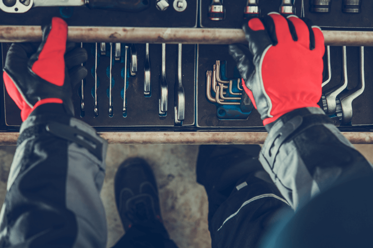 Nahaufnahme von Händen in roten Arbeitshandschuhen, die eine Werkzeuglade mit verschiedenen Werkzeugen wie Schraubenschlüsseln und Inbusschlüsseln durchsuchen.