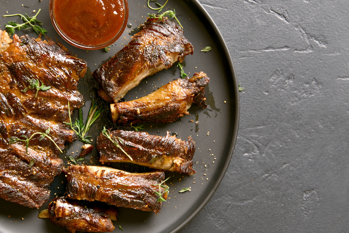Gegrillte Spareribs auf einem schwarzen Teller, garnierte mit Kräutern und einer Schale BBQ-Sauce.