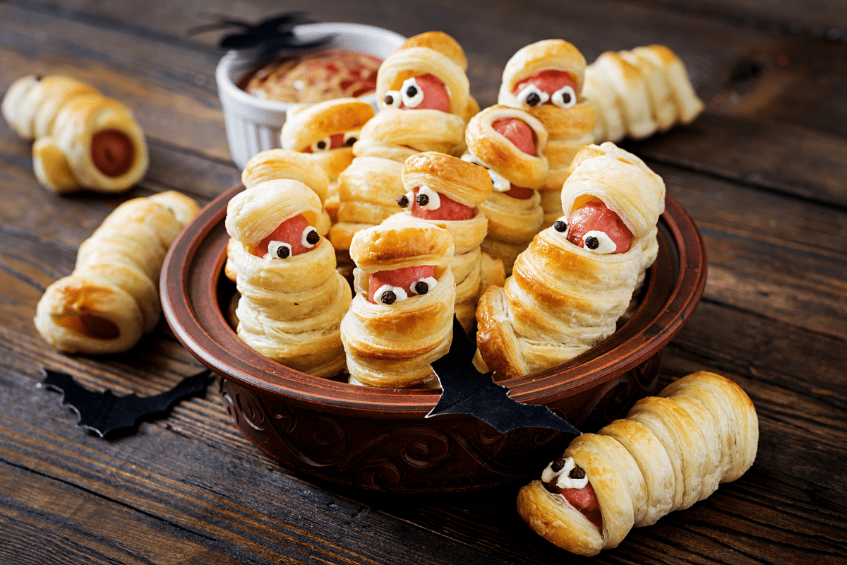Würstchen in Blätterteig, dekoriert als Mumien mit Augen, serviert in einer Schüssel. Perfekt für Halloween-Snacks.