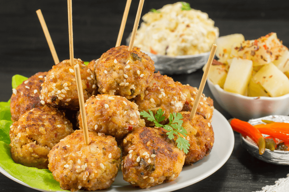 Herzhafte Hackbällchen auf einem Teller, mit Sesam und Spießen, umgeben von Beilagen wie Reis und Gemüse.