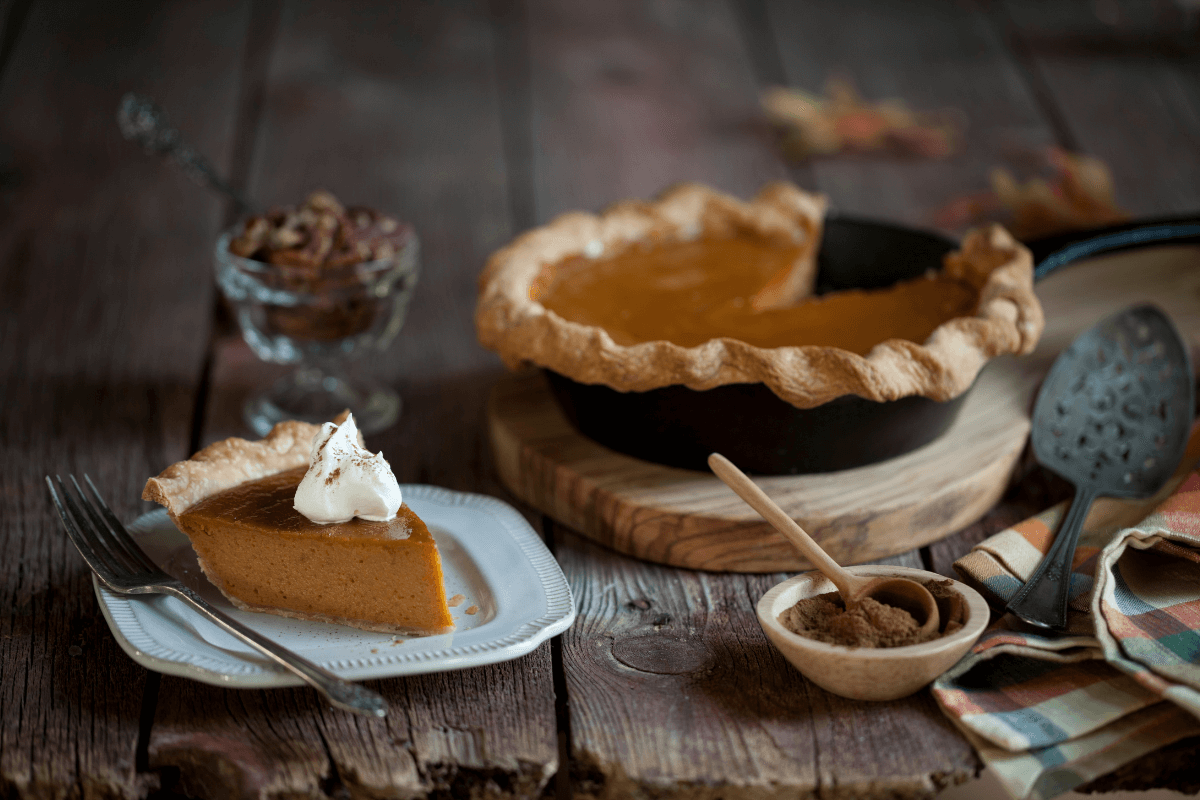 Ein Stück Kürbiskuchen auf einem weißen Teller, dekoriert mit Sahne, daneben eine ganze Torte und Gewürze.