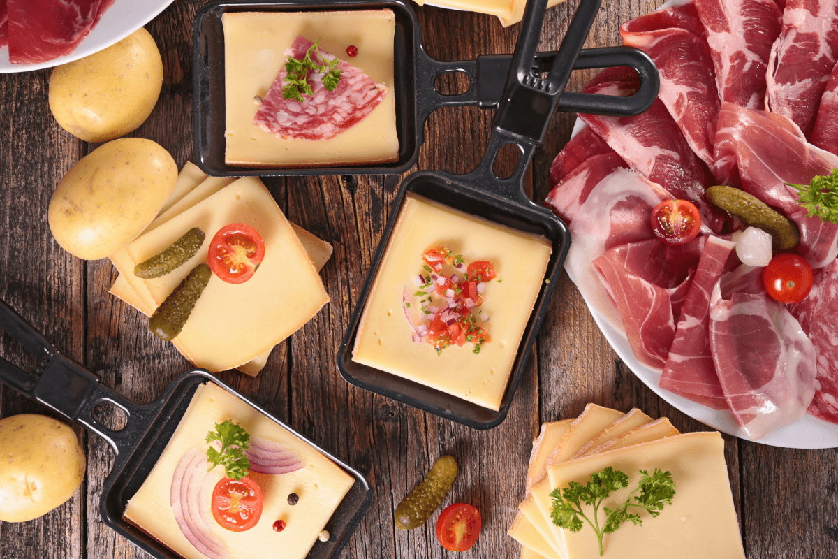 Raclette-Gerichte mit Käse, Schinken, Salami, Kartoffeln, Tomaten und Gewürzgurken auf einem Holztisch.
