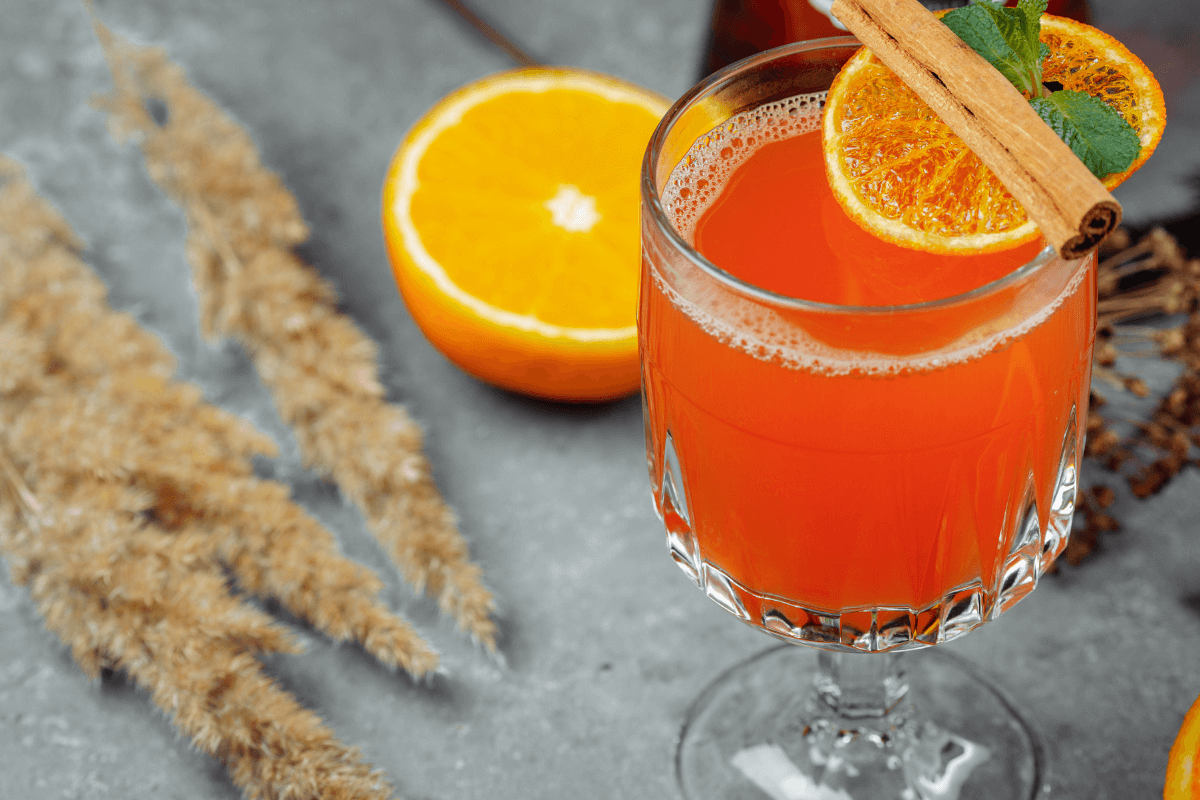 Ein Glas mit Hot Aperol, dekoriert mit einer getrockneten Orangenscheibe, Zimt und frischer Minze. Im Hintergrund eine halbe Orange und dekorative Gräser.