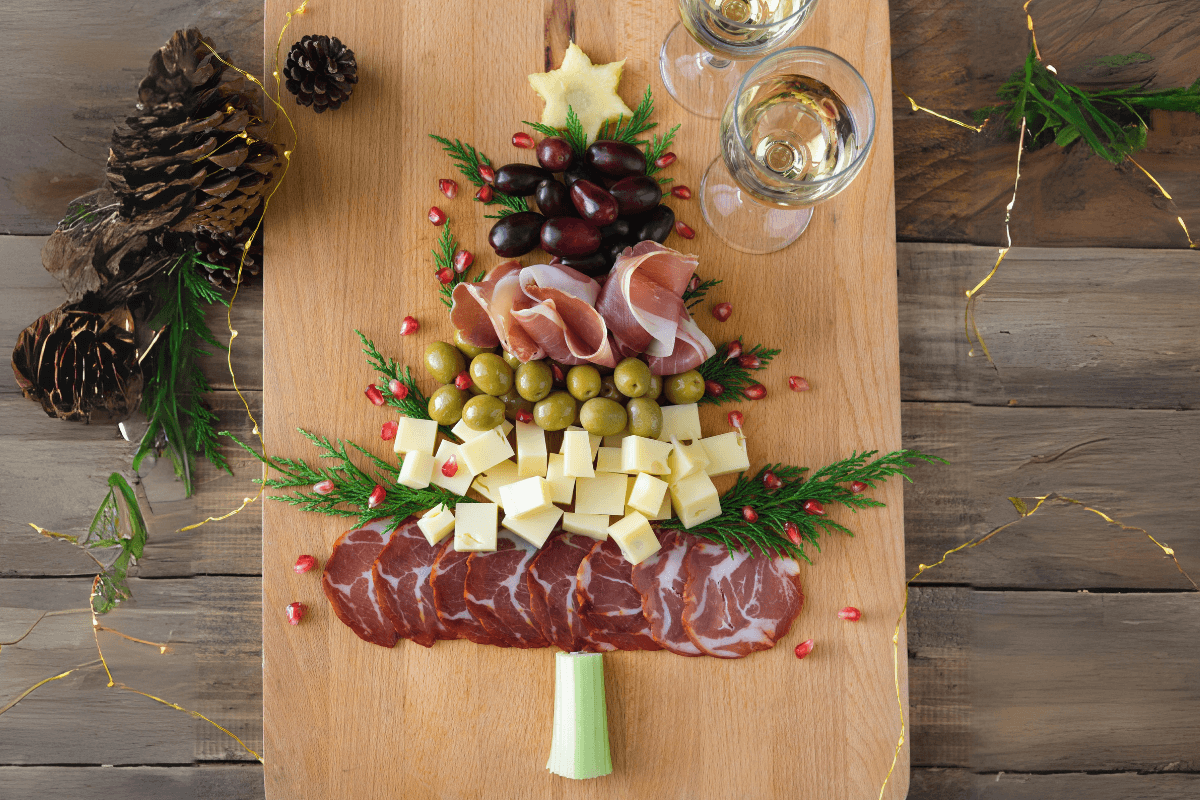 Ein festlich dekoriertes Holzbrett in Form eines Weihnachtsbaums, belegt mit Käse, Oliven, Schinken, Trauben und Dekoration.