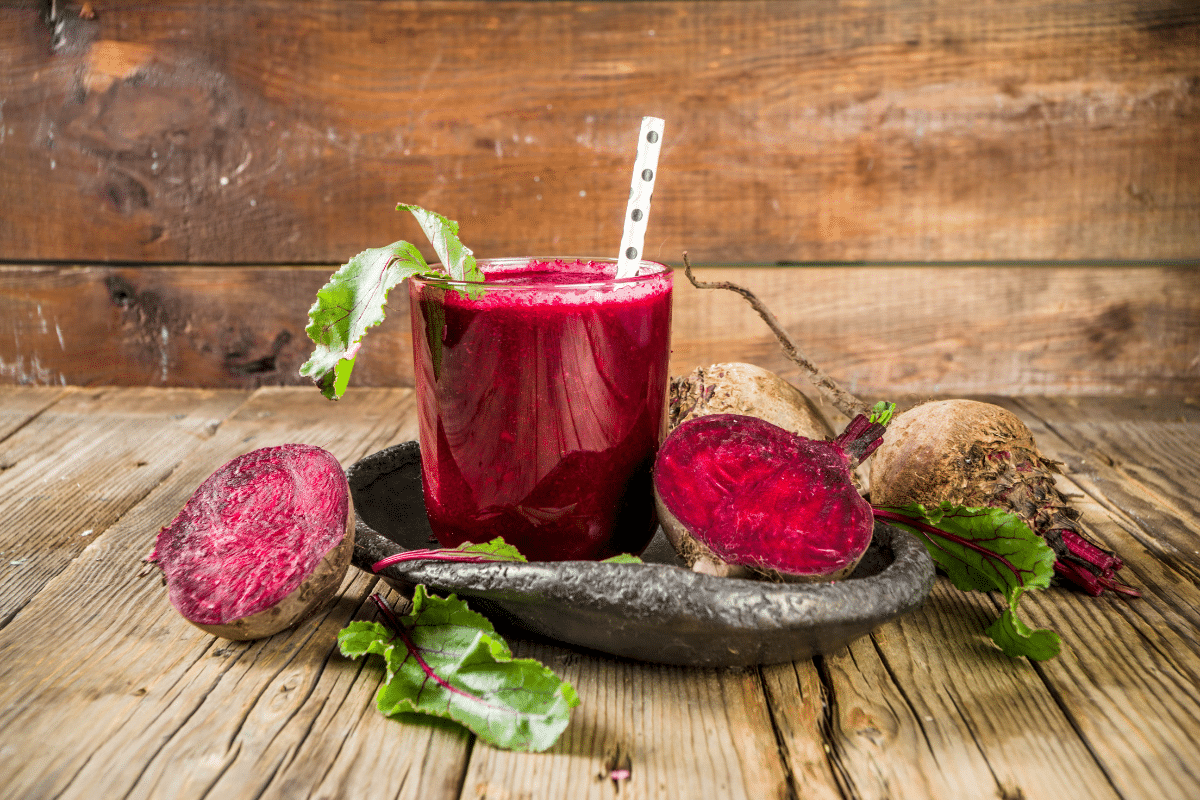 Rote-Bete-Smoothie in einem Glas mit einem Strohhalm, umgeben von frischen Rüben und Blättern auf einem dunklen Teller.