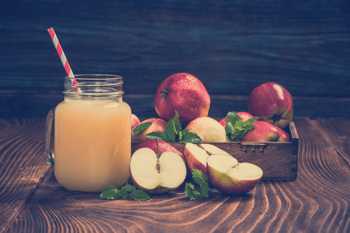 Glas mit Apfelsaft und gestreiftem Strohhalm, umgeben von frischen Äpfeln und Minzblättern auf einem Holztisch.
