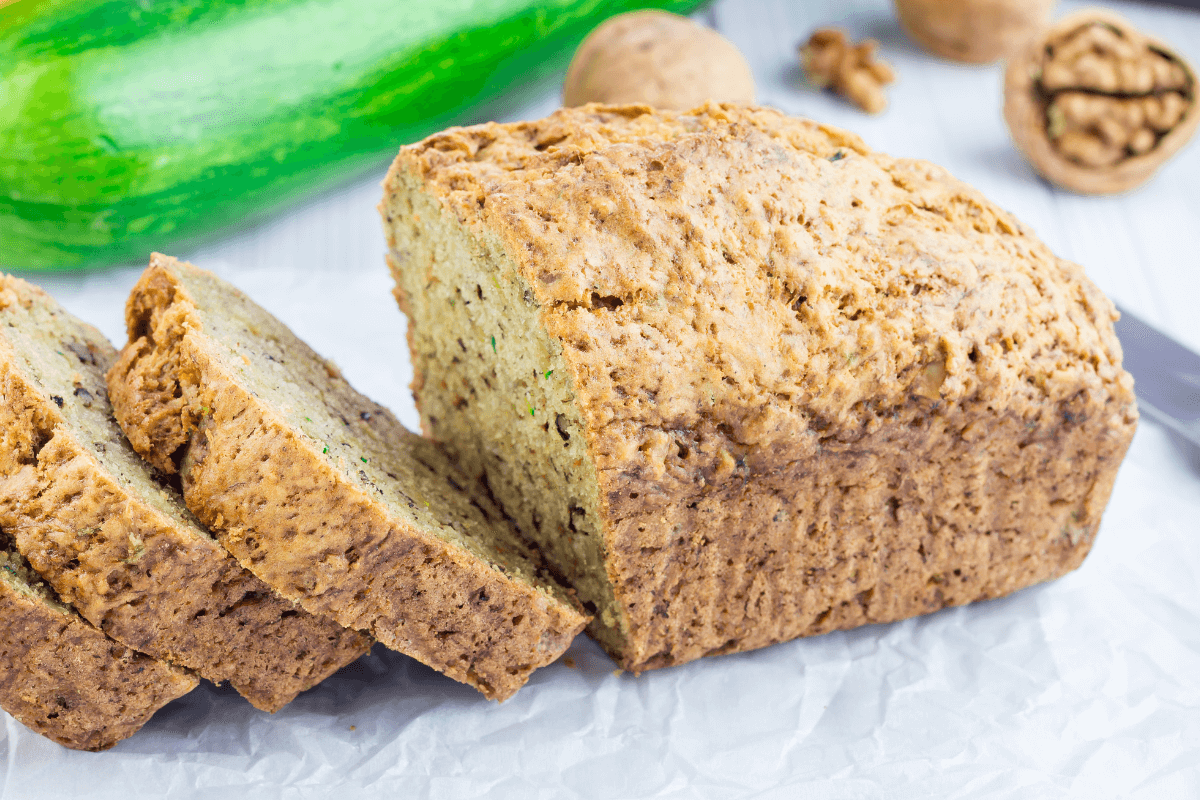 Dinkelbrot mit Zucchini und Walnüssen, in Scheiben geschnitten, auf weißem Papier. Im Hintergrund frische Zucchini und Walnüsse.