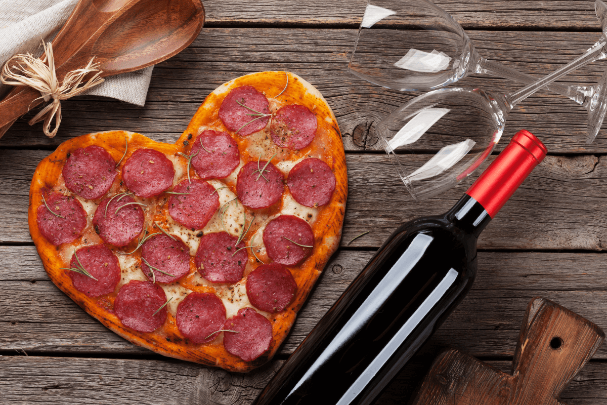 Herzförmige Pizza mit Salami und Kräutern, neben einer Flasche Rotwein und zwei Weingläsern auf einem Holztisch.