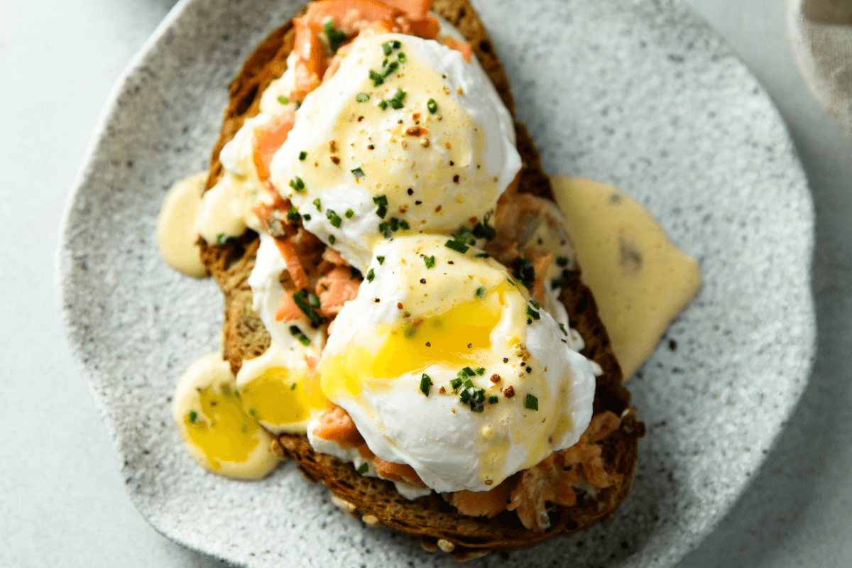 Zwei pochierte Eier auf Vollkornbrot mit Lachs, garniert mit Schnittlauch und Sauce Hollandaise, serviert auf einem grauen Teller.