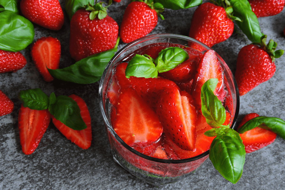 Glas mit Erdbeeren und Basilikumblättern, umgeben von frischen Erdbeeren auf grauem Hintergrund.