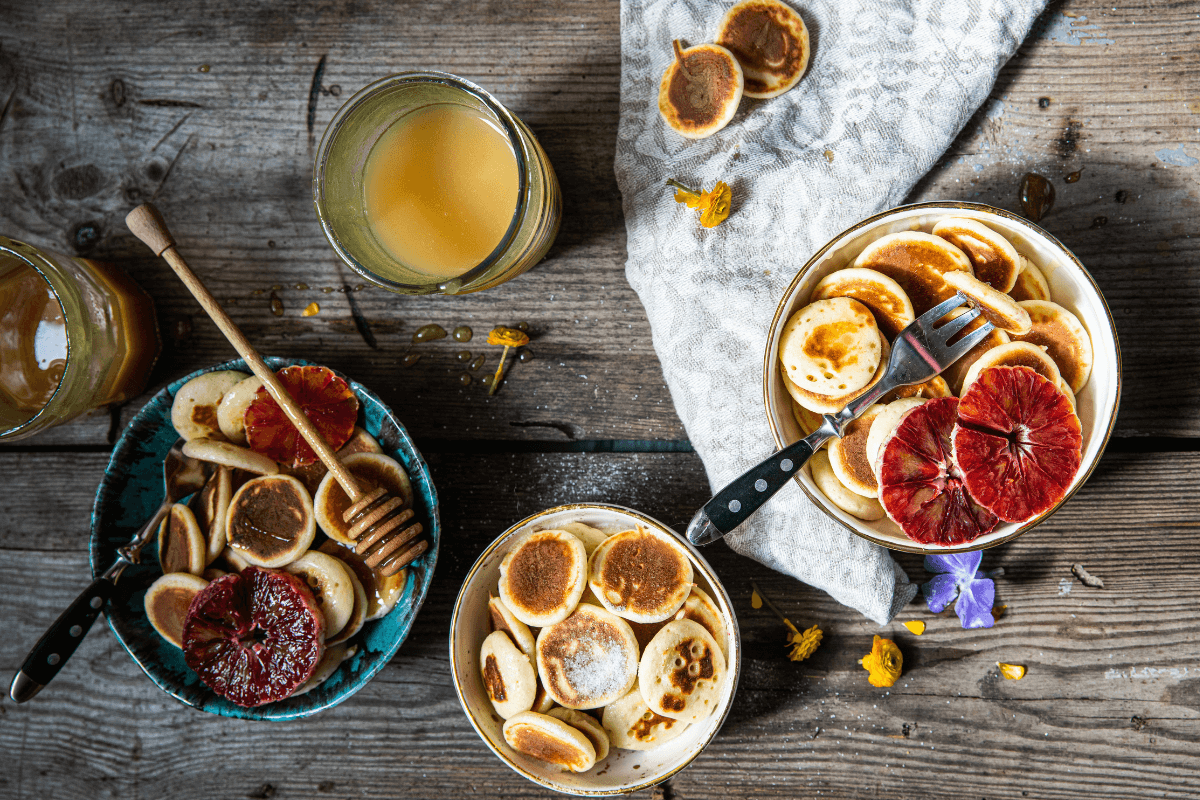 Kleine Pancakes in zwei Schalen, dekoriert mit Blutorangen, Honig und einem Glas Saft auf einem rustikalen Holztisch.