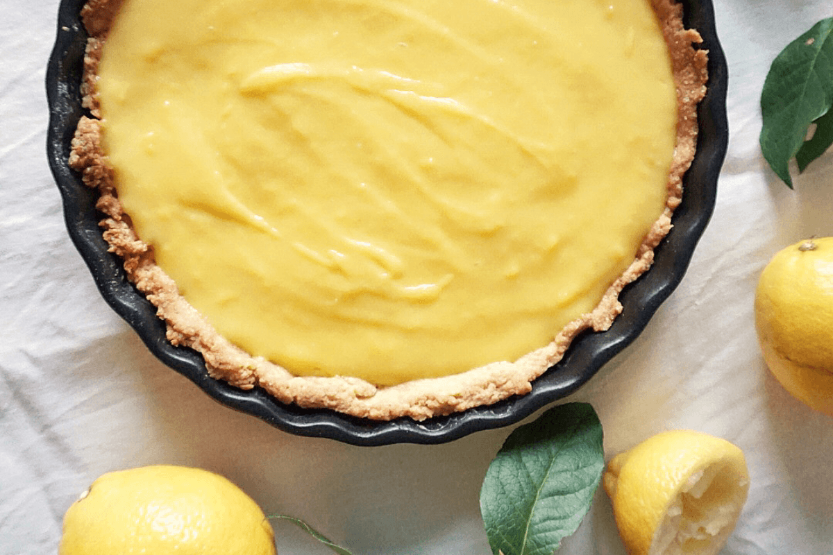 Zitronentarte in einer schwarzen Pieform mit einer glatten, gelben Füllung und einem knusprigen Rand. Frische Zitronen daneben.