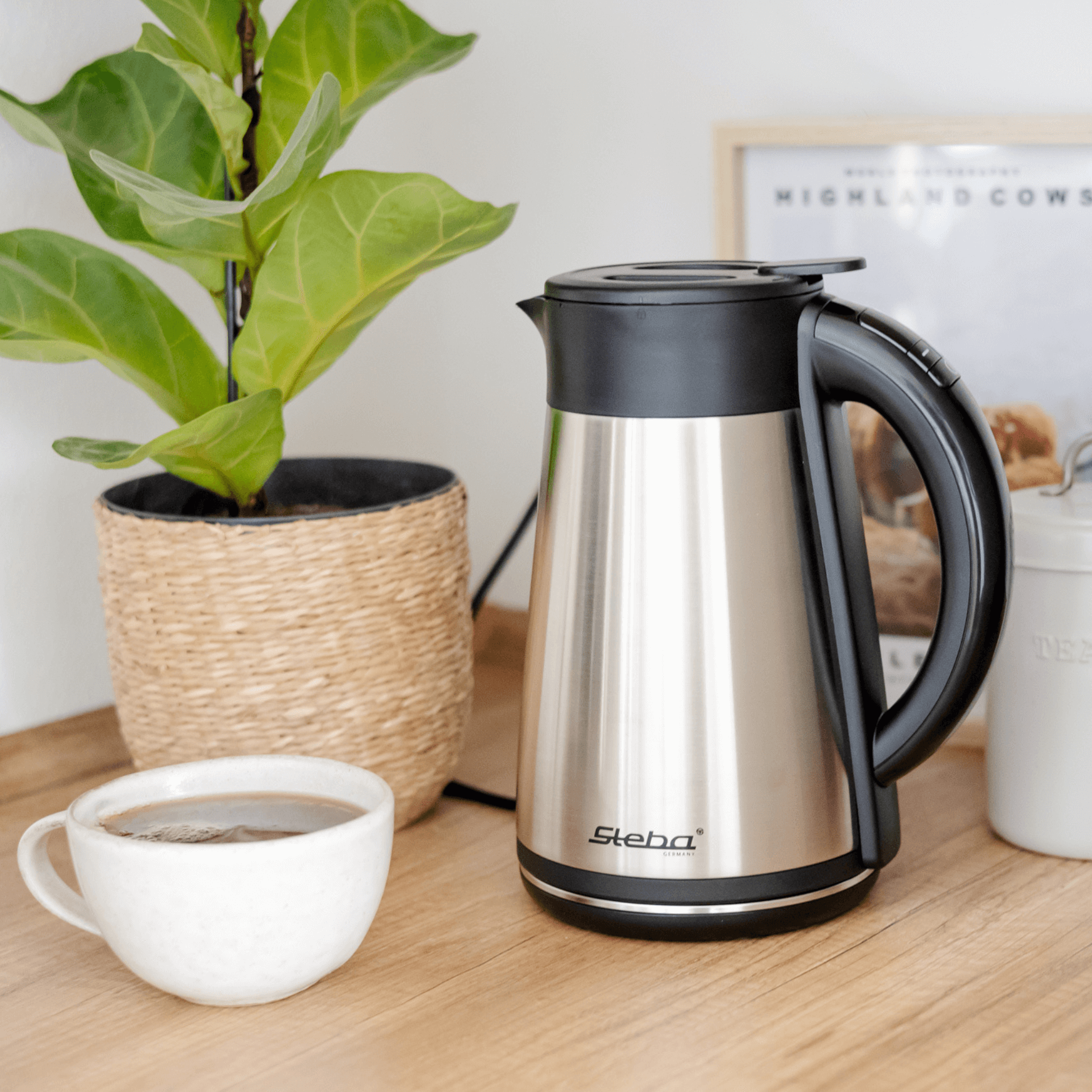 Edelstahl-Wasserkocher WK31THERMO von Steba mit schwarzem Deckel und Griff, neben einer Tasse Kaffee und einer Pflanze.