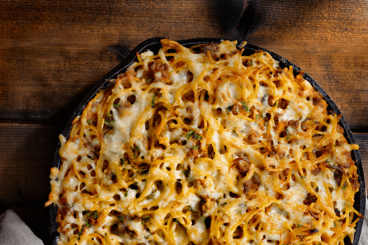 Überbackene Spaghetti mit Käse und Fleisch, serviert in einer schwarzen Pfanne auf einem Holzuntergrund.