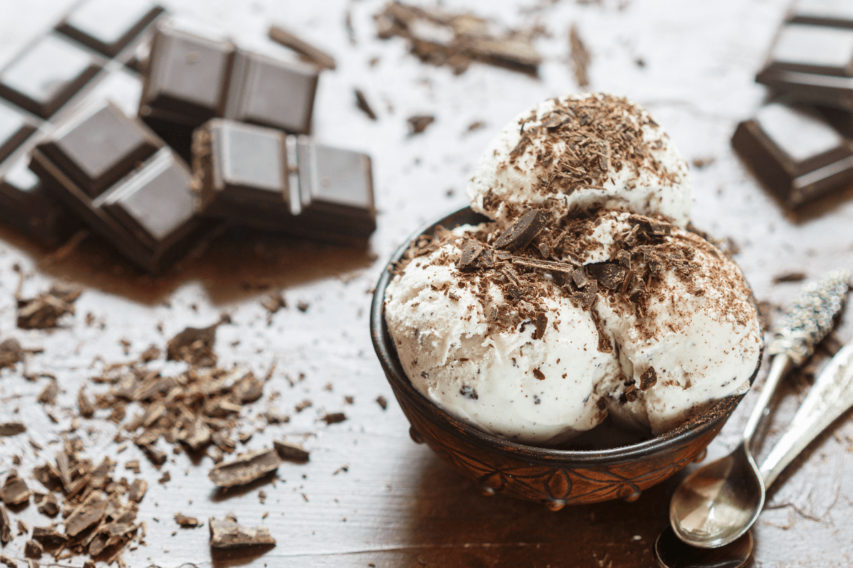 Schüssel mit Stracciatella-Eis, dekoriert mit Schokoladenraspeln, umgeben von Schokoladentafeln und Kakaobruch.