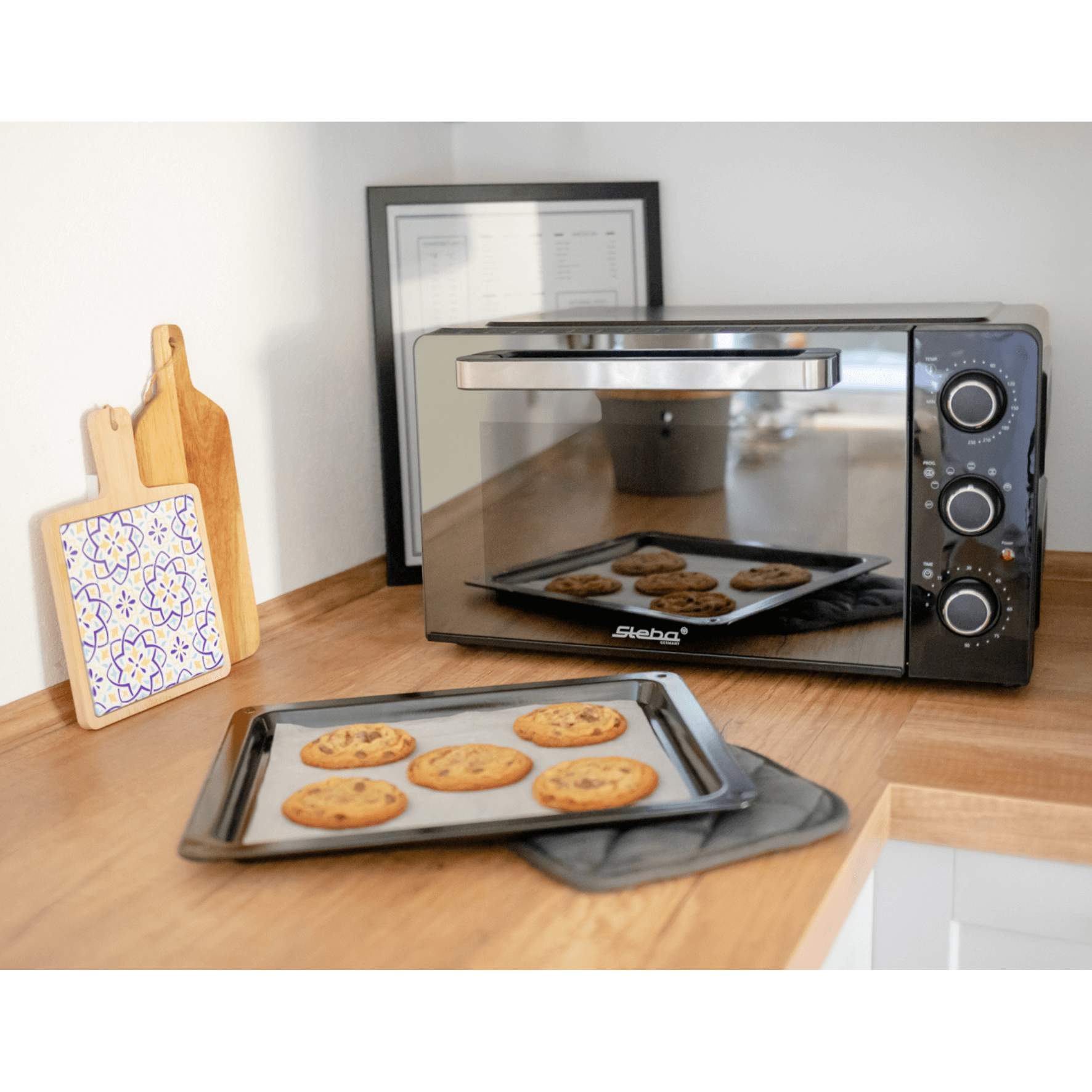 Steba Grillbackofen auf einer Holzarbeitsfläche mit Keksen auf einem Blech. Dekorative Holzbrettchen im Hintergrund.