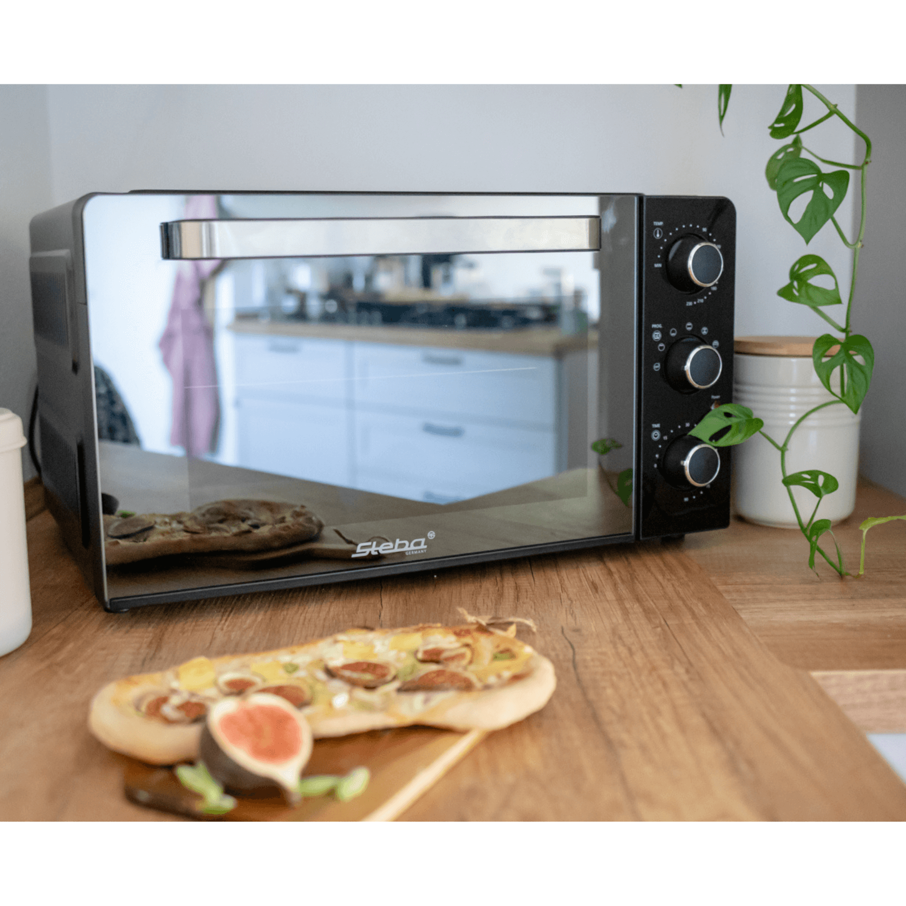 Steba Grillbackofen KB224 mit reflektierender Front und drei Drehreglern, platziert auf einem Holzsortiment neben einem belegten Brot.