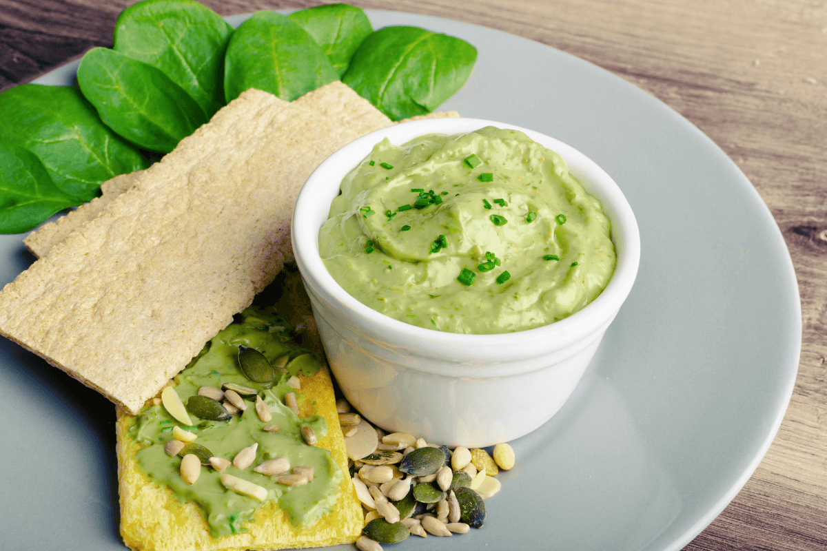 Schüssel mit Green Goddess Dip, umgeben von Crackern und frischem Spinat auf einem grauen Teller.